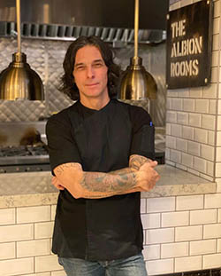 Chef Rene Rodriguez leans against a counter with his arms folded, the Albion Rooms sign visible over his left shoulder.
